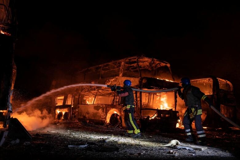 Firefighters hose down a fire, at night, in front of the burning remains of two buses. 