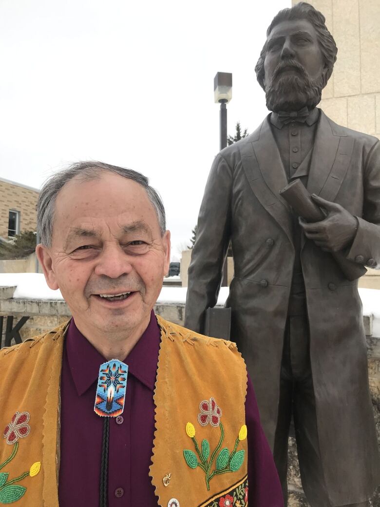 An Indigenous man stands in front of a statue, smiling.