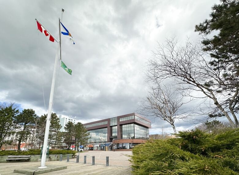 A large brick building with lots of windows is seen in the distance through some trees on the right and a flag pole on the left with the Canadian, Nova Scotian and CBRM flags fluttering in the breeze.