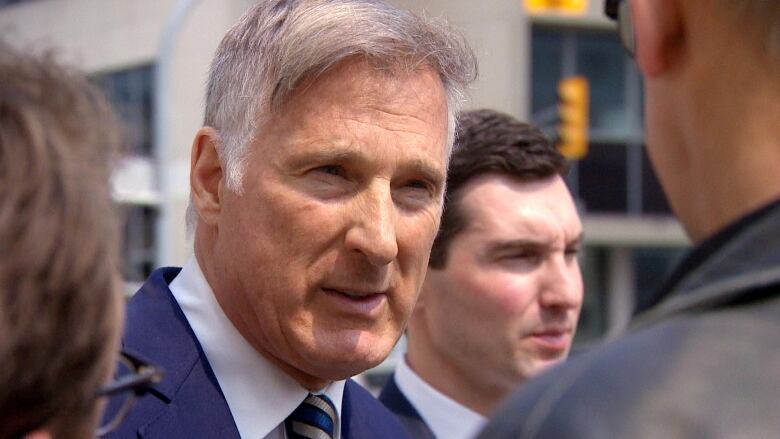 A man with short grey hair and wearing a navy suit and tie speaks outside.