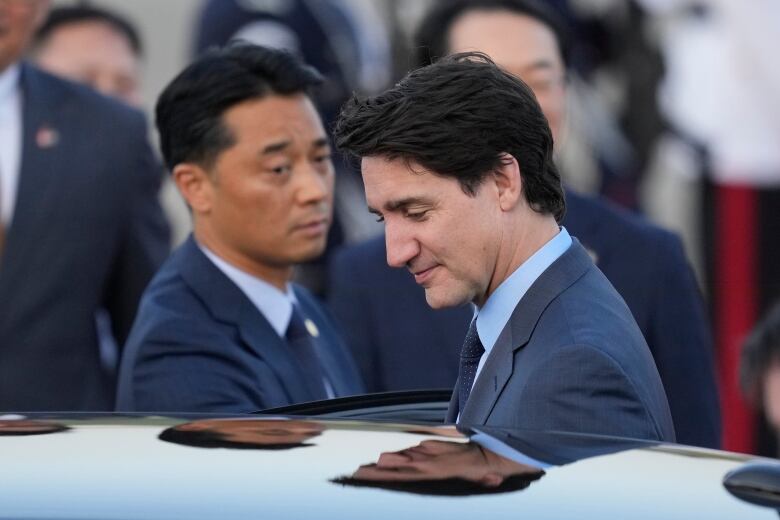 A man wearing a suit opens a car door to get into the vehicle, as another man in a suit looks on in the background.