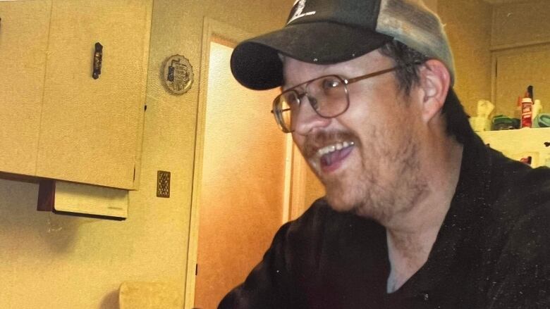 A man in aviator glasses, a black shirt and a ball cap smiles at someone off camera.