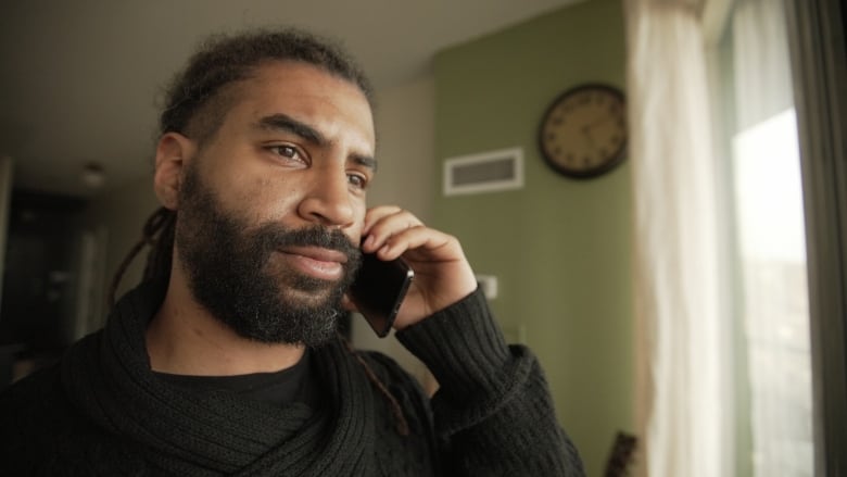 A man with a full beard wearing a black sweater holds a mobile phone to his ear while looking off camera.