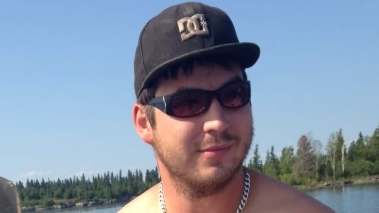 A shirtless man in sunglasses and a baseball cap is seen near water.