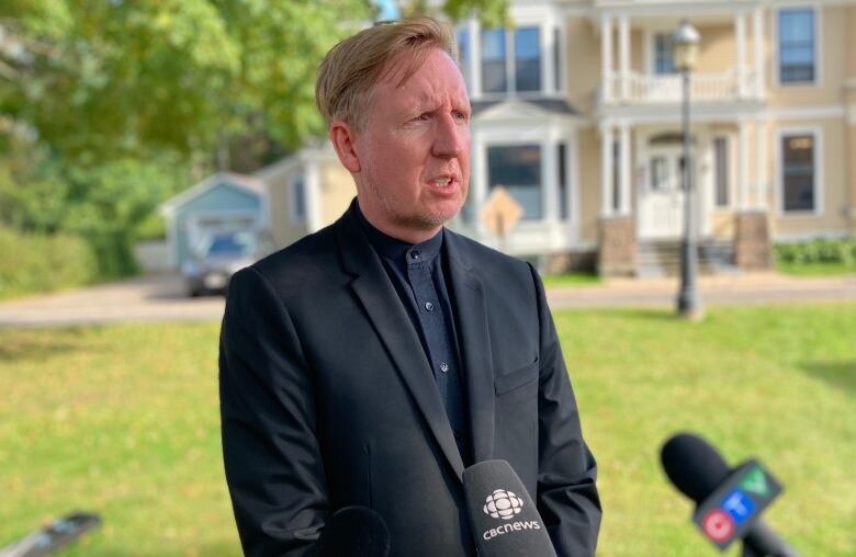 A man with blond hair in a dark suit stands outside talking to reporters.