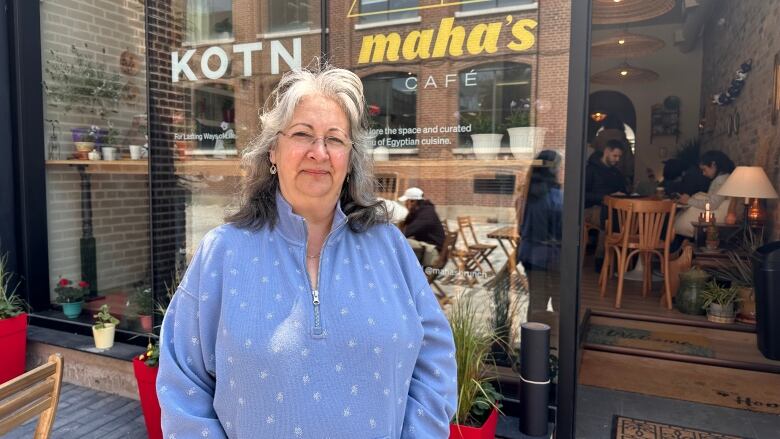 A woman stands in front of a storefront, she's wearing a blue quarter zip and has grey hair.