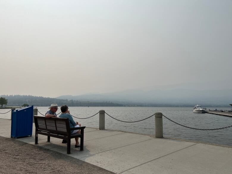 Two people sit on a bench overlooking a lake with smoky skies.