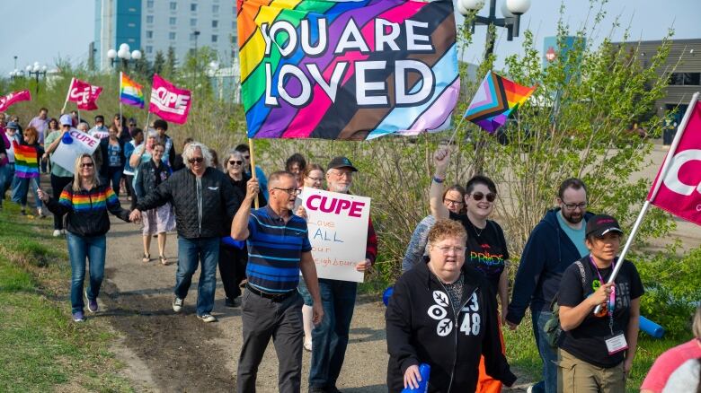 A group of around 80 people march carrying Pride and CUPE flags in support of the LGBTQ community.