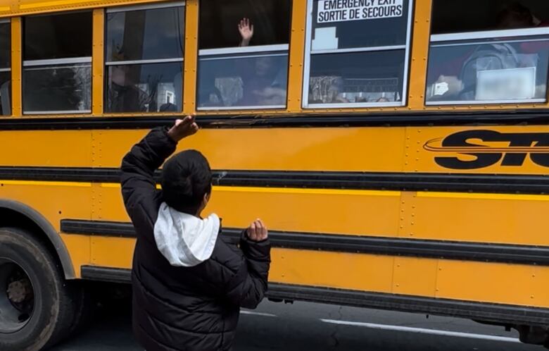 A boy stands with his back to the camera, waving goodbye to a friend on a school bus.