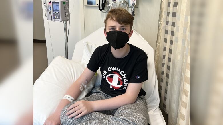 A boy can be seen wearing a black tshirt and a mask while sitting on a hospital bed.