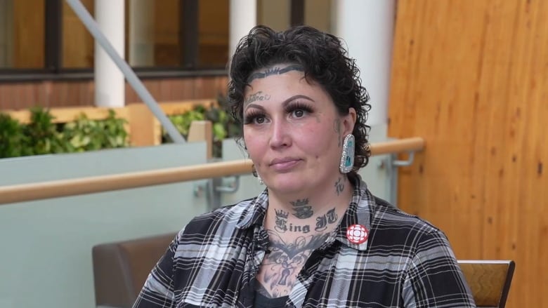 A woman with tattoos and short, curly black hair is pictured wearing a black and grey plaid shirt during an interview.