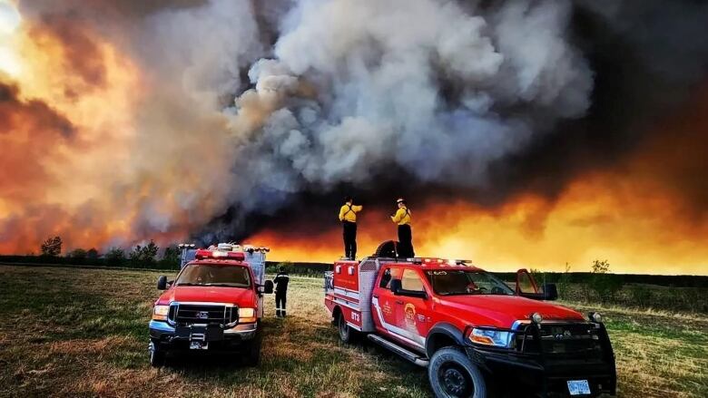 Firefighters standing on the back of red firetrucks look back at a sky full of flames and smoke in this surreal wide-angle photo.