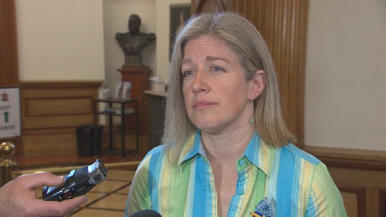 a woman with light hair and a blue and yellow striped shirt talks to reporters.