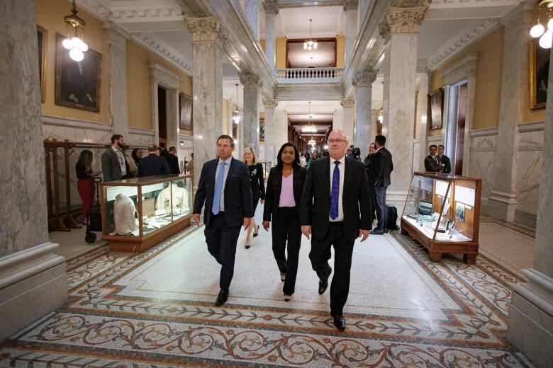 Three people walk down an ornate hallway with marble columns and tiled flooring.