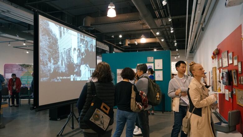 Visitors to the museum take a look at Lee's exhibition. Seen here is a white screen, with an image of a family projected onto it, and multiple people milling about and looking at artworks and artifacts.