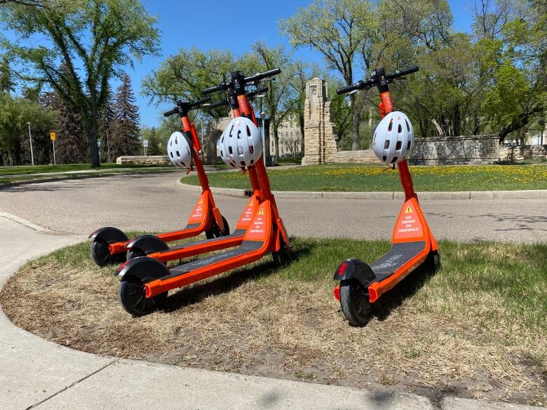 Four bright orange e-scooters are sitting on the grass with helmets hanging from the handle bars.