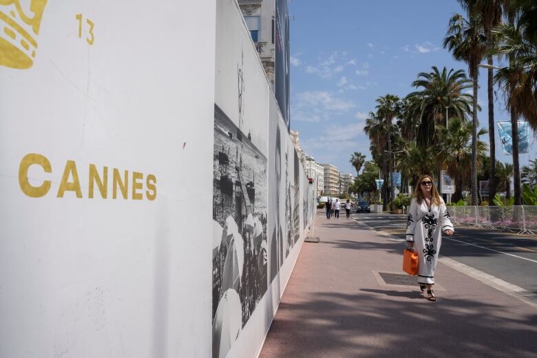 A woman walks down a palm tree-lined street with posters nearby advertising Cannes.