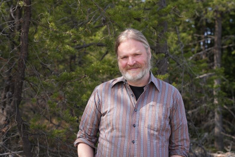 A man poses with a forest background. He has grey and light brown hair tied back and a beard. He wears a plaid button up collar shirt. 