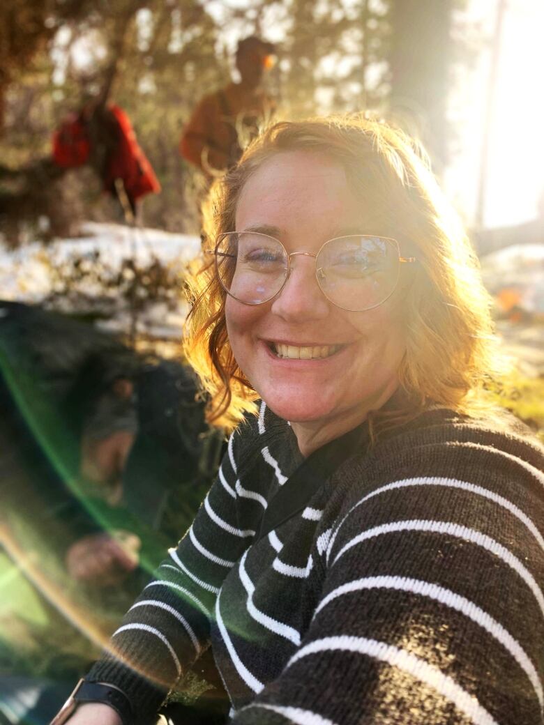 A picture of a woman smiling with brown shoulder length hair, wearing glasses and a black shirt with thin white stripes. The sun shines bright behind her, creating a glow around her.  