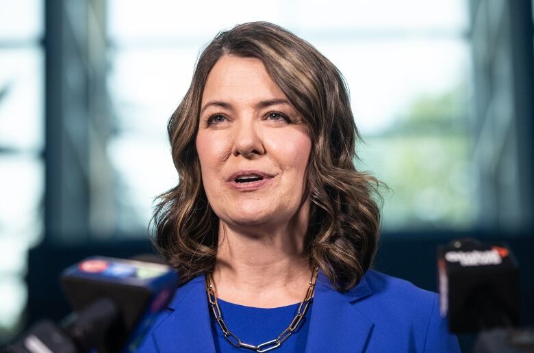 Danielle Smith, wearing a royal blue blazer and matching shell and a gold link necklace looks up as she speaks into the microphone in this closeup photo.