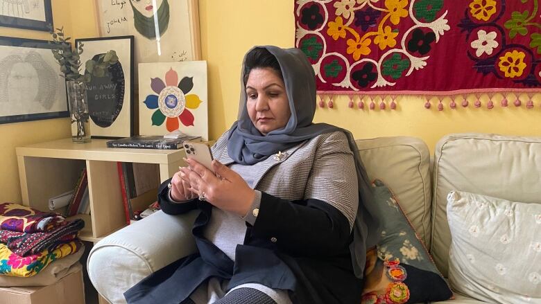 A woman in a headscarf sits on a couch, checking her phone.