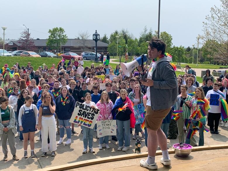 A student with short, dark hair and wearing a gray hoodie and brown shorts, holds  a megaphone from a stage in the foreground, looking out at a crowd of a couple of hundred students. One student in the front row is holding a home-made poster that says Policy 713 saves lives.