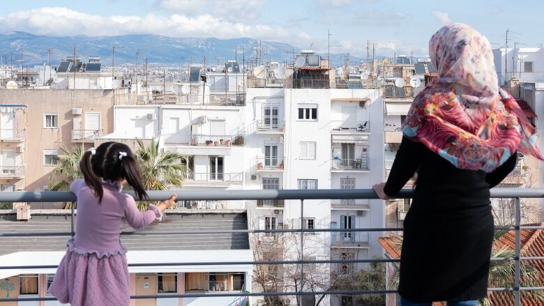 A woman in a head scarf and a child with pigtails look over a railing into a lowrise cityscape with mountains in the background.