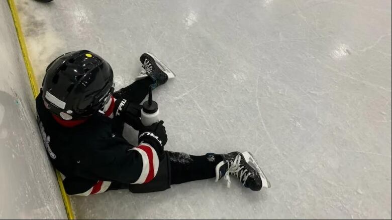 A child hockey player sits on the ice with his back against the boards.