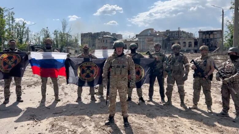 A man in military fatigues stands in front of others similarly dressed, some holding flags including a Russian flag.