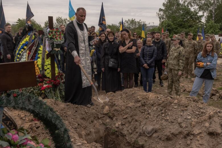 Mourners watch a man in religious vestments throw a shovelful of dirt into an open grave.