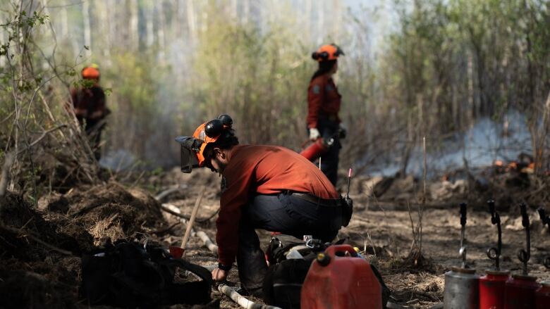 Three firefighters work as fires burn in the background in shrubbery.