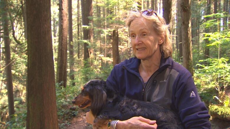 A woman holds a Dachsund in her arms on a forested trail.