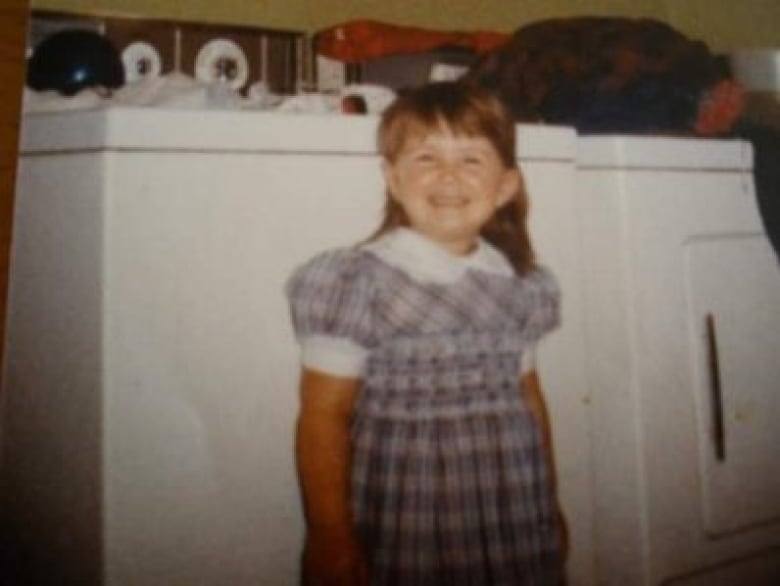 An old colour photograph of a young, smiling girl with red hair. She's wearing a blue and purple plaid dress with puffy short sleeves.