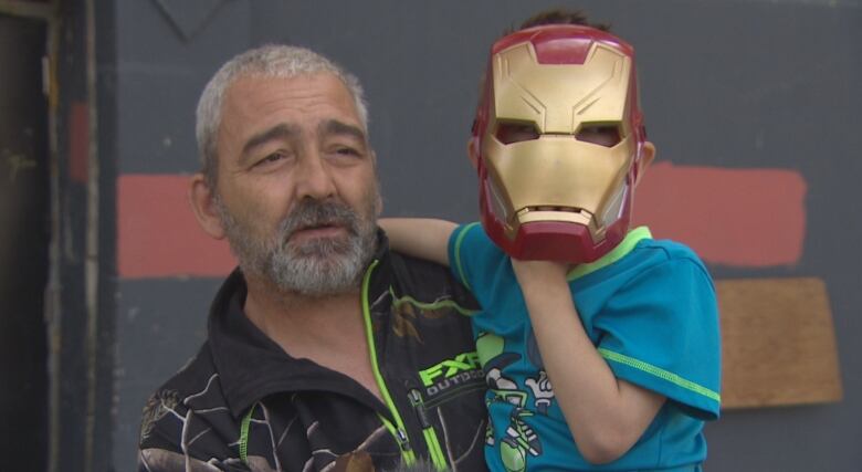 A man in short grey hair and a grey beard holds a child who is wearing an Iron Man Halloween mask.