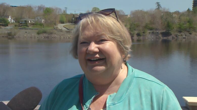 A white woman with a round face and short blonde hair, wearing a turqoise cardigan, stands on a deck overlooking a lake. Trees and homes on the far shore are in the background