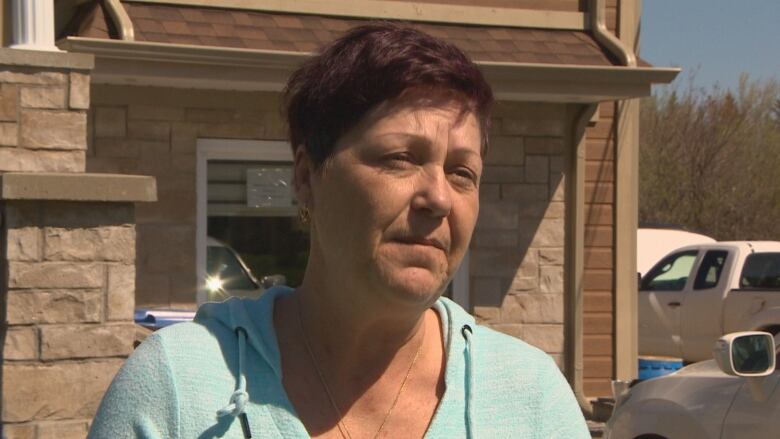 A middle aged woman with short auburn hair gives an interview in front of a home with vinyl and stone siding.