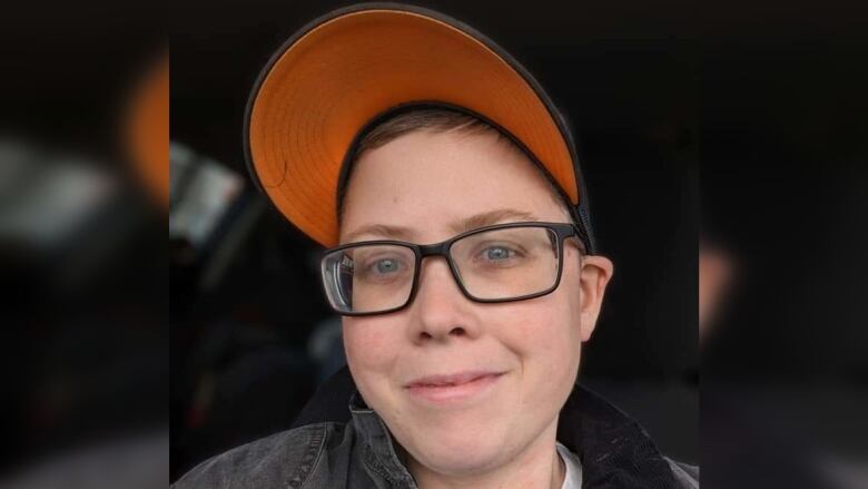 A head and shoulders shot of a person in a baseball cap and black-rimmed glasses. 