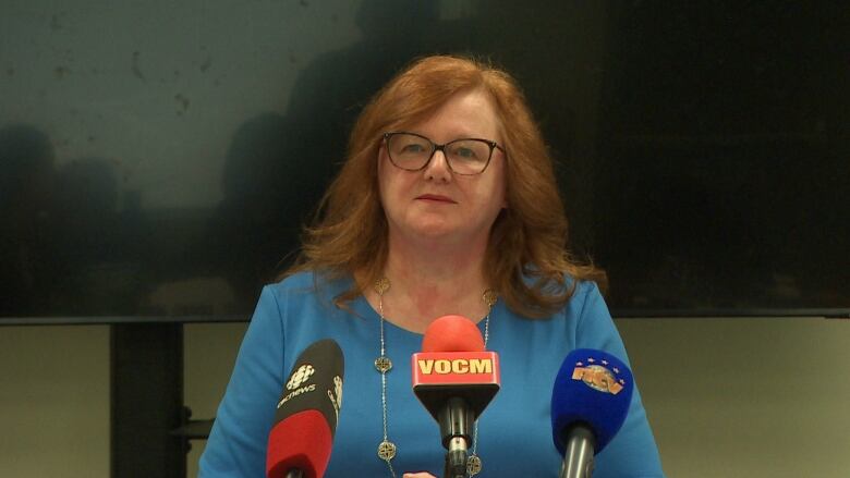 A woman with long, red hair sits in front of microphones in a board room.