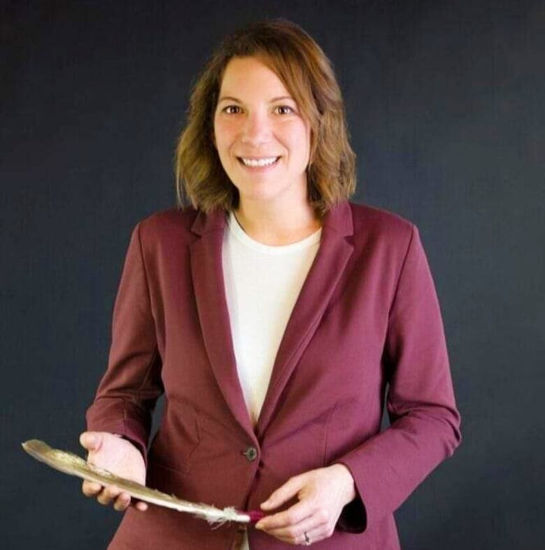 A woman with shoulder-length brown hair wearing a red blazer and holding an eagle feather