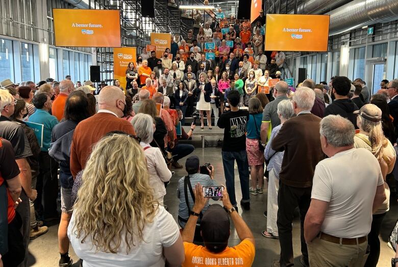 Several people watch Rachel Notley speak, as several more people stand behind her on a cascade of risers.