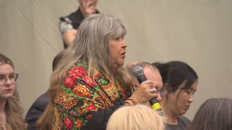 A woman with long grey hair in a flowery shawl stands among a group of people with a microphone in her hand.