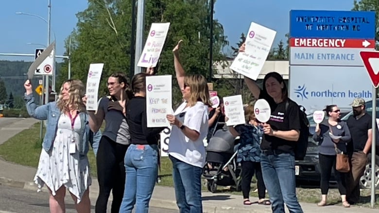 A group of people hold signs up outside by a sign that says Emergency. Main Entrance.