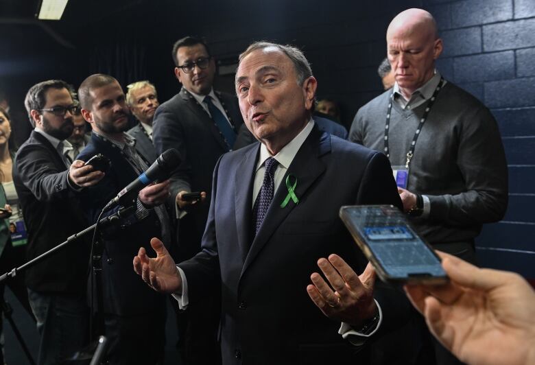 A man wearing a navy blue suit speaks to reporters clustered around him, with microphones and recording devices stretched out.