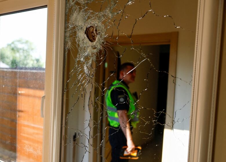 A closeup of a hole in a window, with cracked glass, is shown. Inside the building, a police officer stands in a doorway.