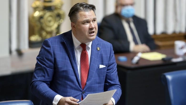 A man with dark hair in a blue checked suit and a red tie holds a paper and speaks in a legislative chamber. 