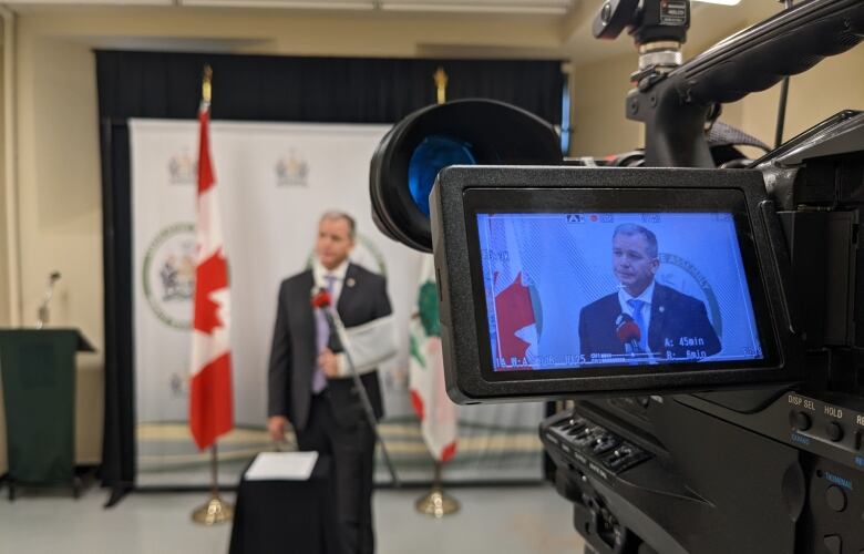 Man in business suit stands in front of television camera.