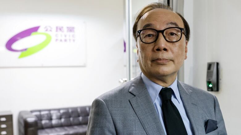 A man wearing a suit and tie stands in a room as a sign with Chinese writing is seen behind him.