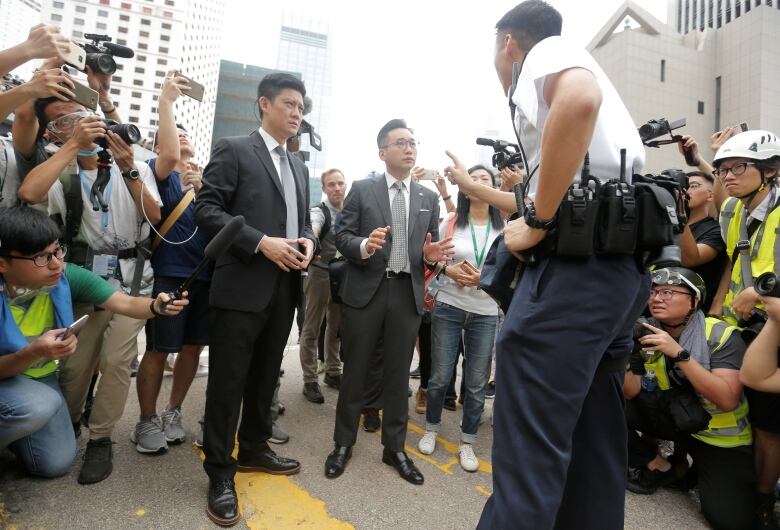 Two men speak with a police officer as people with tape recorders and cameras surround them.