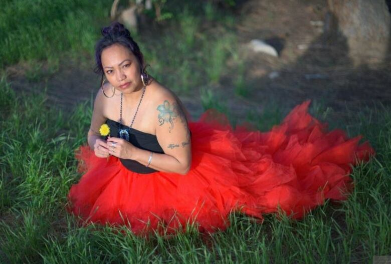 A woman wearing a black and red flowing dress poses in green vegetation while holding a yellow flower.