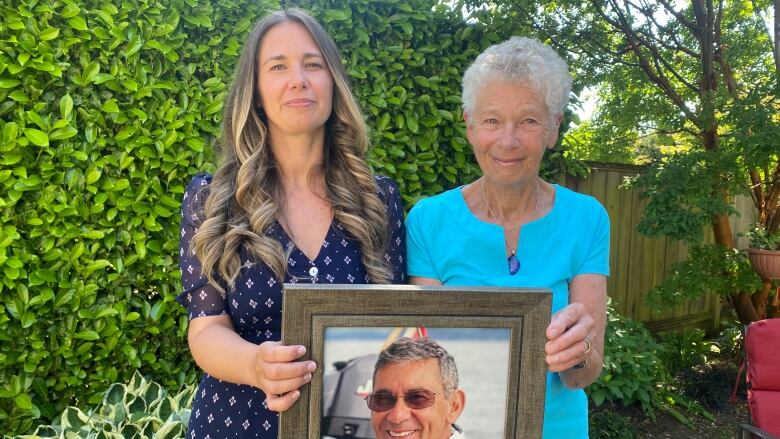 Two women hold a picture of an older man in a yard.